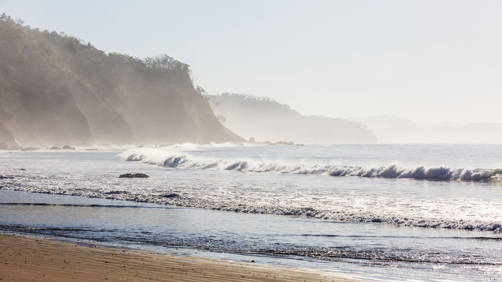 Waves gently crash on a sandy beach along Costa Rica's Pacific coast, with mist-covered cliffs in the background under a hazy sky.